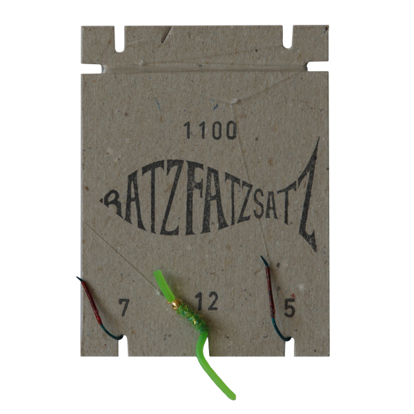 Auf einem quadratischen Karton sind in Fischform „BATZFATZSATZ“ sowie die Nummern 7, 12 und 5 abgebildet, dazu drei Haken – einer davon mit einem Grün-Köder – in Anlehnung an den RatzFatzSatz 1100 (Grün).