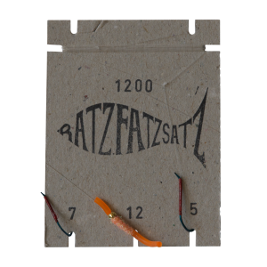 Auf einem Stück Pappe mit der Aufschrift „RatzFatzSatz 1200 (Orange)“ befinden sich drei Angelhaken mit den Nummern 7, 12 und 5. An dem Haken mit der Nummer 12 ist ein orangefarbener und ein kupferfarbener Köder befestigt.