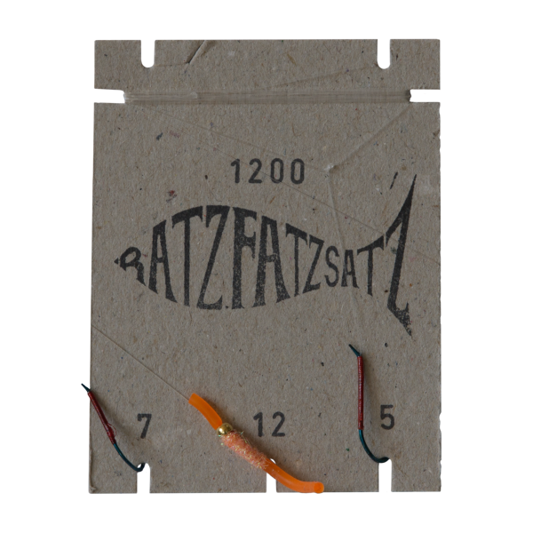 Auf einem Stück Pappe mit der Aufschrift „RatzFatzSatz 1200 (Orange)“ befinden sich drei Angelhaken mit den Nummern 7, 12 und 5. An dem Haken mit der Nummer 12 ist ein orangefarbener und ein kupferfarbener Köder befestigt.