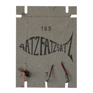 Der RatzFatzSatz 189 (Orange & Rot) besteht aus einer rechteckigen Karte in Fischform, den Zahlen 189 und 79 in Orange sowie zwei Angelhaken und einem mit Schnur befestigten Fliegenköder.