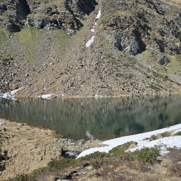 Der Lagh de Calvaresc, ein Bergsee mit spiegelglatter Oberfläche, umgeben von steilen Felswänden und Schneeresten.