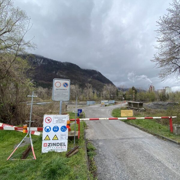 Eine schmale Straße zum Caluori Weiher ist durch eine rot-weiße Barriere mit mehreren Warn- und Hinweisschildern gesperrt, umgeben von Bäumen und Gras, mit Bergen und bewölktem Himmel im Hintergrund.