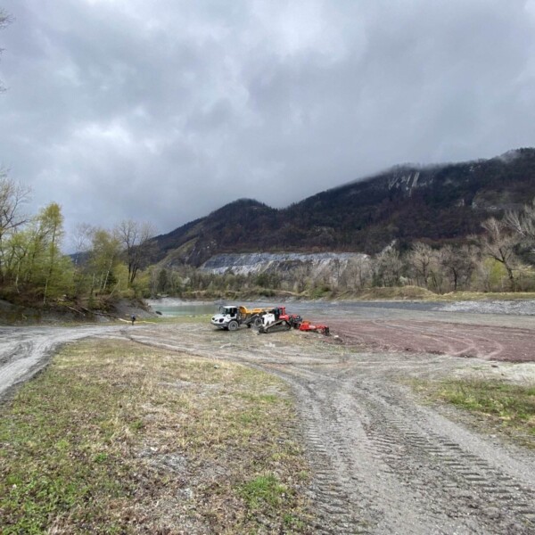 Baufahrzeuge parken auf einer weiten, schlammigen Lichtung in der Nähe des Caluori Weiher, umgeben von Bäumen und Bergen unter einem bewölkten Himmel. Reifenspuren zeichnen den Boden, und etwas Grün säumt das Gebiet.