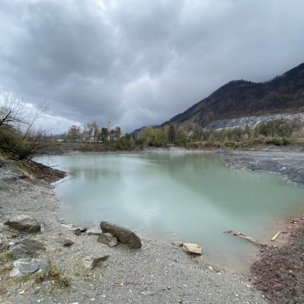 Ein wolkiger Himmel ragt über den Caluori Weiher, einen kleinen türkisfarbenen Teich, umgeben von felsigem, kargem Boden und spärlichen Bäumen, mit bewaldeten Hügeln und Bergen im Hintergrund.