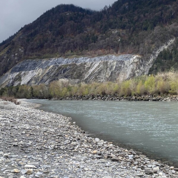Ein flaches, felsiges Flussufer führt zu einem breiten, ruhigen Fluss. Am gegenüberliegenden Ufer befinden sich grüne Bäume und ein steiler, felsiger Hang mit freiliegenden Steinschichten unter einem bewölkten Himmel.