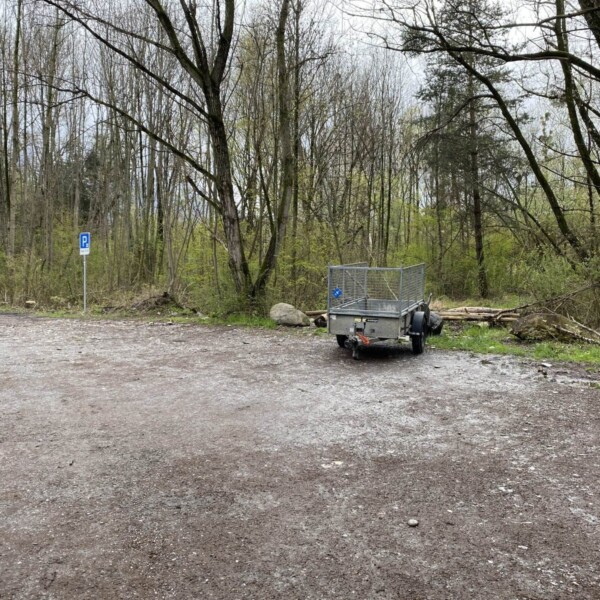 Ein kleiner Metallanhänger steht auf einem Schotterplatz in der Nähe eines blauen Parkschildes am Zizerser Weiher, umgeben von blattlosen Bäumen und grünem Unterholz in einem Waldgebiet.