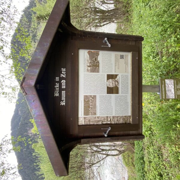 Eine hölzerne Außenanzeigetafel mit dem Titel „Blicke in Raum und Zeit“ mit Text, Fotos und einem QR-Code steht auf einem Metallmast inmitten des Grüns des Biotops Salischinas, im Hintergrund sind Bäume und Hügel zu sehen.