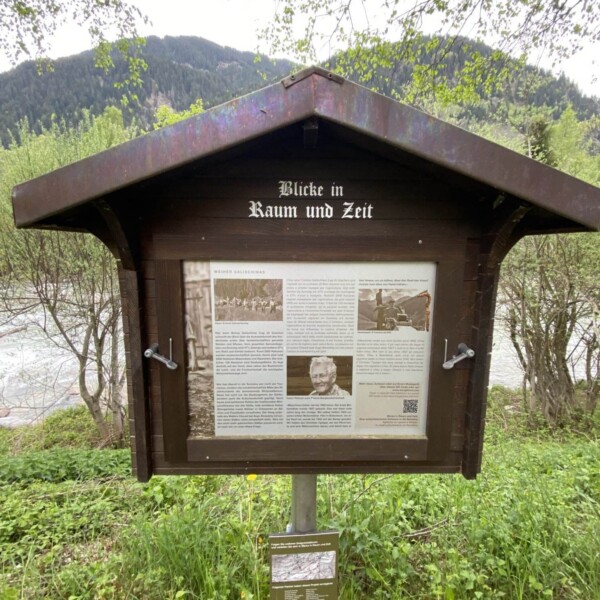 Eine hölzerne Außenanzeigetafel mit dem Titel „Blicke in Raum und Zeit“ mit Text, Fotos und einem QR-Code steht auf einem Metallmast inmitten des Grüns des Biotops Salischinas, im Hintergrund sind Bäume und Hügel zu sehen.