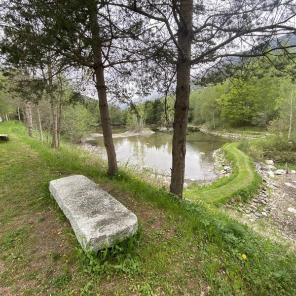 Eine Steinbank steht auf Grasboden neben einem kleinen Teich im üppigen Biotop von Salischinas, umgeben von Bäumen und Felsen. In der Ferne ist entlang des Weges eine weitere Bank zu sehen.
