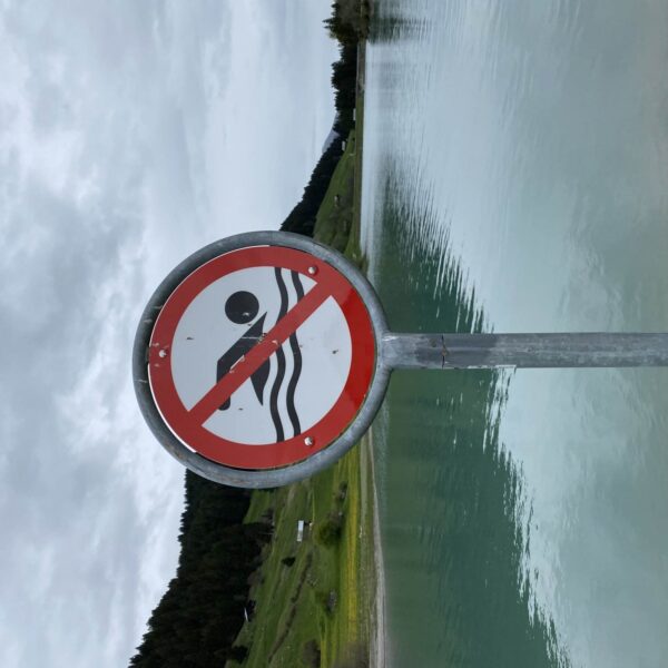 Am ruhigen Brigelsersee (Lag da Breil) steht ein rundes Schild mit einer roten diagonalen Linie, die ein Badesymbol durchkreuzt. Es weist darauf hin, dass das Baden verboten ist. Im Hintergrund sind grüne Hügel und ein bewölkter Himmel zu sehen.