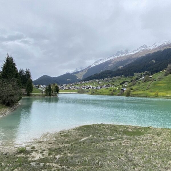 Ein ruhiger türkisfarbener See, bekannt als Brigelsersee oder Lag da Breil, mit grasbewachsenen und von Bäumen gesäumten Ufern liegt vor einem kleinen Dorf, eingerahmt von bewaldeten Hügeln und schneebedeckten Bergen unter einem bewölkten Himmel.