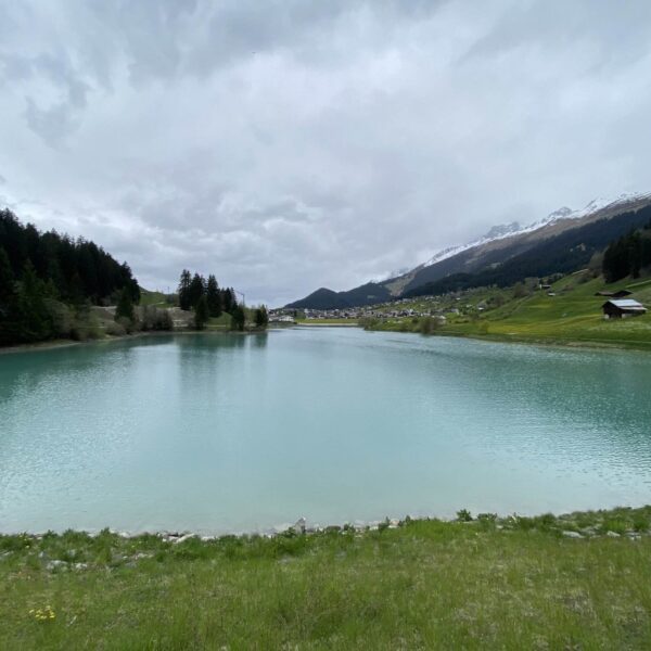Ein türkisfarbener See, bekannt als Brigelsersee oder Lag da Breil, ist von grünem Gras und Bäumen umgeben, mit ein paar kleinen Häusern, die den rechten Hang sprenkeln, und Bergen, die sich im Hintergrund unter einem bewölkten Himmel erheben.