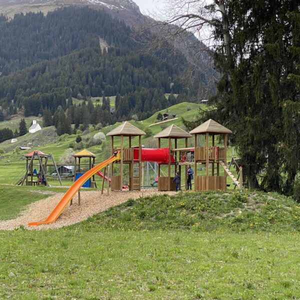 Ein Holzspielplatz mit Rutschen, Brücken und Türmen steht auf einer Wiese in der Nähe des Brigelsersees, umgeben von Hügeln, Bäumen und Bergen. Kinder spielen unter einem bewölkten Himmel auf den Geräten.