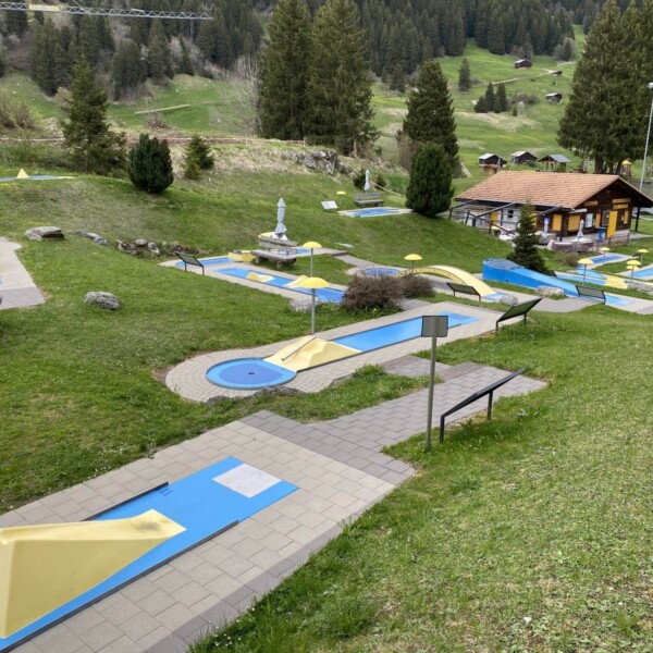 Minigolfplatz im Freien mit blauen und gelben Hindernissen in der Nähe des Lag da Breil, umgeben von grünem Gras, Bäumen und einem Holzgebäude im Hintergrund. Bänke und ein Spielplatz sind ebenfalls zu sehen.