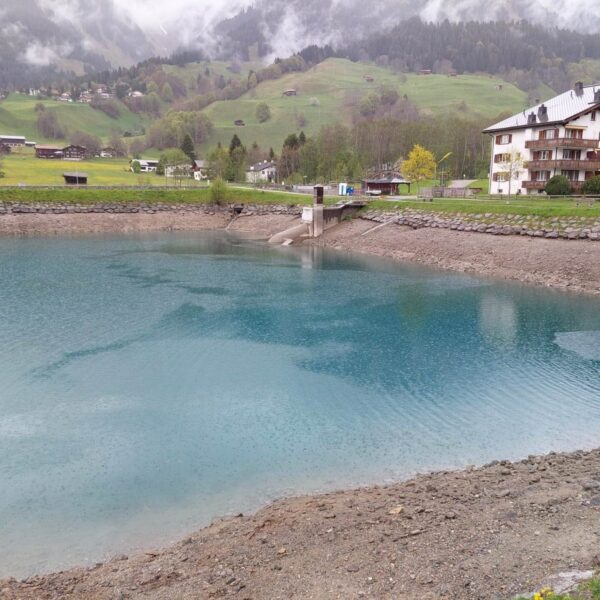 Ein kleiner türkisfarbener See, der Doggilochsee, mit niedrigem Wasserstand ist umgeben von grünen Hügeln, verstreuten Häusern und einem mehrstöckigen Chalet unter bewölktem, nebligem Himmel in einer bergigen Landschaft.