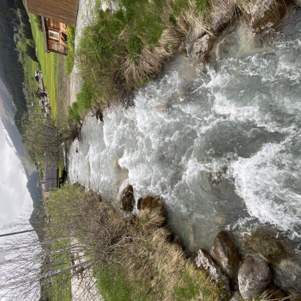 Ein schnell fließender Bach fließt durch grasbewachsene Ufer mit Felsen in Flem, umgeben von Bergen und verstreuten Häusern unter einem bewölkten Brigels-Himmel.