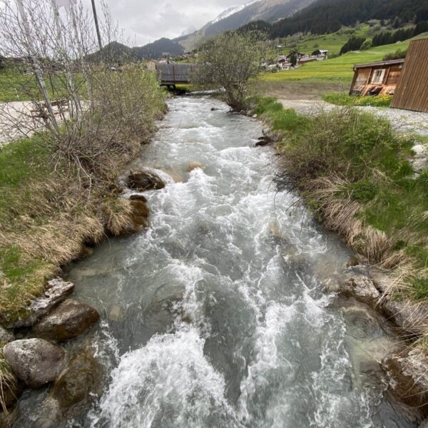 Ein schnell fließender Bach fließt durch grasbewachsene Ufer mit Felsen in Flem, umgeben von Bergen und verstreuten Häusern unter einem bewölkten Brigels-Himmel.
