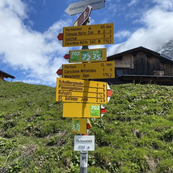 Wegweiser auf einem grasbewachsenen Hang mit Wanderanweisungen und -zeiten zu verschiedenen Zielen, darunter Partnunsee, Sulzfluh und St. Antönier Joch. Im Hintergrund sind ein Holzgebäude und ein blauer Himmel mit Wolken zu sehen.