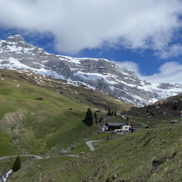 Eine malerische Aussicht auf schneebedeckte Berge unter einem teilweise bewölkten Himmel, mit grünen Hügeln und einer kurvenreichen Straße, die zu einem kleinen Haus führt, das im Tal darunter liegt.