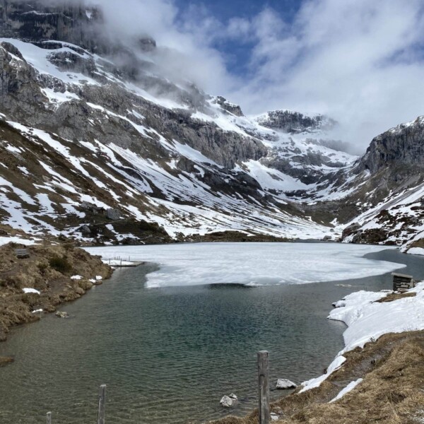 Ein teilweise zugefrorener Bergsee, umgeben von felsigen, schneebedeckten Hängen unter einem überwiegend bewölkten Himmel, mit Grasflecken und schmelzendem Schnee entlang des Ufers.