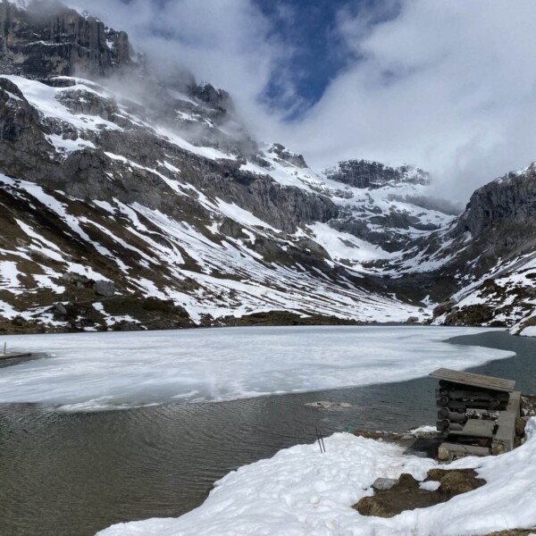 Ein teilweise zugefrorener Bergsee, umgeben von schneebedeckten Felshängen unter einem bewölkten blauen Himmel, mit einer kleinen Steinstruktur nahe der Wasserkante im Vordergrund.