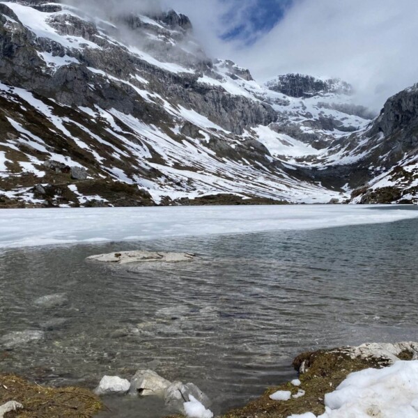 Ein teilweise zugefrorener Bergsee mit schneebedeckten Felshängen und Gipfeln im Hintergrund unter einem bewölkten Himmel. Das Wasser ist in Ufernähe klar und ruhig, am Rand liegen Schneeflecken.