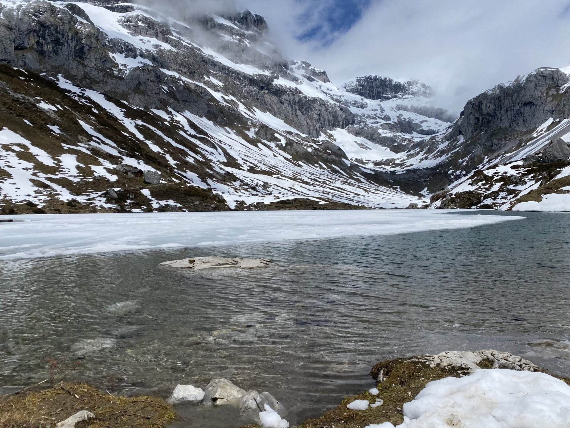 Ein teilweise zugefrorener Bergsee mit schneebedeckten Felshängen und Gipfeln im Hintergrund unter einem bewölkten Himmel. Das Wasser ist in Ufernähe klar und ruhig, am Rand liegen Schneeflecken.