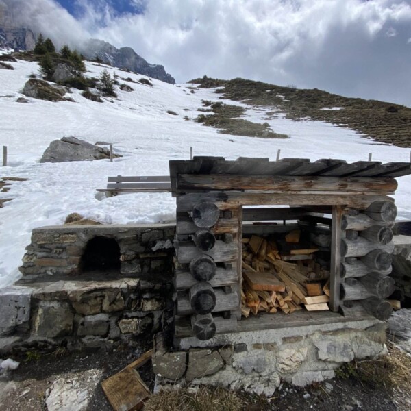 An einem schneebedeckten Berghang steht ein rustikaler Unterstand aus Holz und Stein. Darin stapelt sich Brennholz. Schnee bedeckt den Boden und die Hügel. Im Hintergrund sind unter einem bewölkten Himmel einige Bäume und Felsklippen zu sehen.