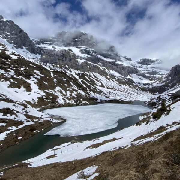 Ein teilweise zugefrorener Alpensee ist von schneebedeckten Bergen und Flecken braunen Grases unter einem bewölkten Himmel umgeben.