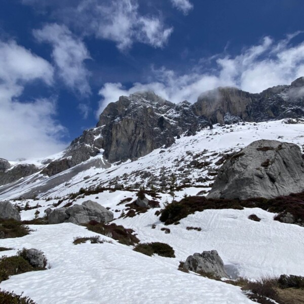 Schneebedeckte Berghänge mit Felsvorsprüngen und Strauchbeständen unter einem teilweise bewölkten blauen Himmel. Einige Bereiche des Berges sind in Nebel oder tief hängende Wolken gehüllt.