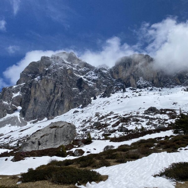 Unter einem teilweise bewölkten blauen Himmel erhebt sich ein schneebedeckter, felsiger Berggipfel, an den unteren Hängen sind Schneeflecken und vereinzelte Sträucher und Bäume zu sehen.