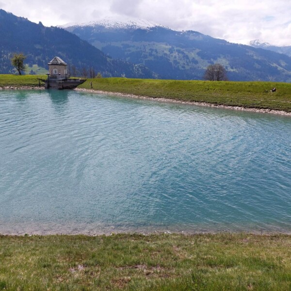 Ein kleiner, ruhiger See mit klarem, blauem Wasser, bekannt als Plävigginersee, ist von grasbewachsenen Ufern und Bergen umgeben. Am Ufer steht eine malerische Holzhütte, und auf den fernen Berggipfeln sind Schneeflecken zu sehen.