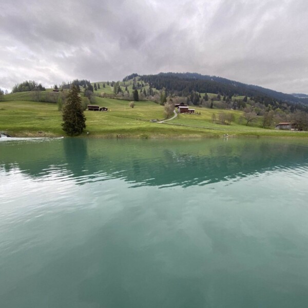 Ein ruhiger, türkisfarbener Plävigginersee spiegelt den bewölkten Himmel wider, mit sanften grünen Hügeln, vereinzelten Bäumen und ein paar rustikalen Häusern im Hintergrund unter einem bedeckten Himmel.