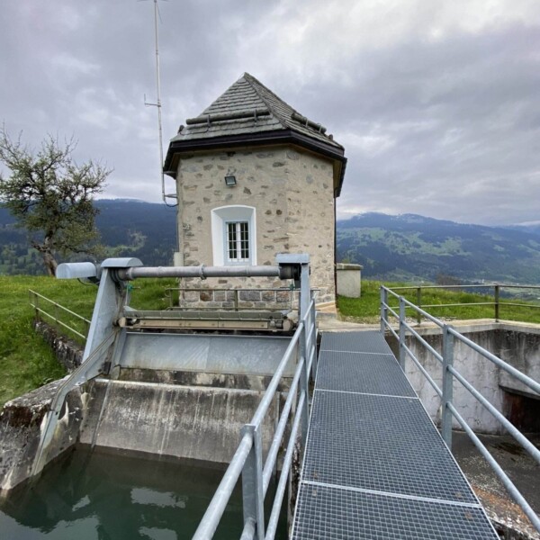 Ein kleines Steingebäude mit Ziegeldach steht am Ende eines Metallstegs über einem Wasserkanal mit Blick auf den Plävigginersee und ist von Grasfeldern und Hügeln unter einem bewölkten Himmel umgeben.