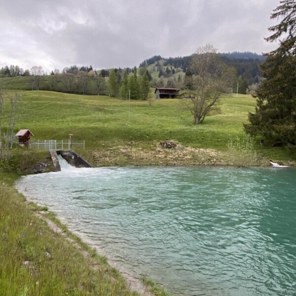 Ein türkisfarbener Fluss fließt durch eine grasbewachsene Hügellandschaft in der Nähe des Plävigginersees. Am Überlauf befindet sich eine kleine Holzhütte. Grüne Bäume, Hütten in der Ferne und ein bewölkter Himmel erstrecken sich über den sanften Hügeln im Hintergrund.