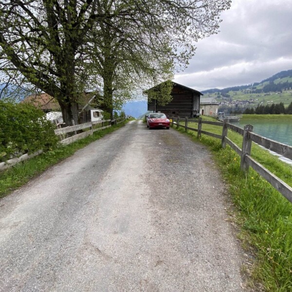 Eine schmale, von Holzzäunen gesäumte Landstraße führt an grünem Gras und Bäumen vorbei zu einem roten Auto in der Nähe von Scheunen am ruhigen, türkisfarbenen Plävigginersee, mit Bergen und bewölktem Himmel im Hintergrund.
