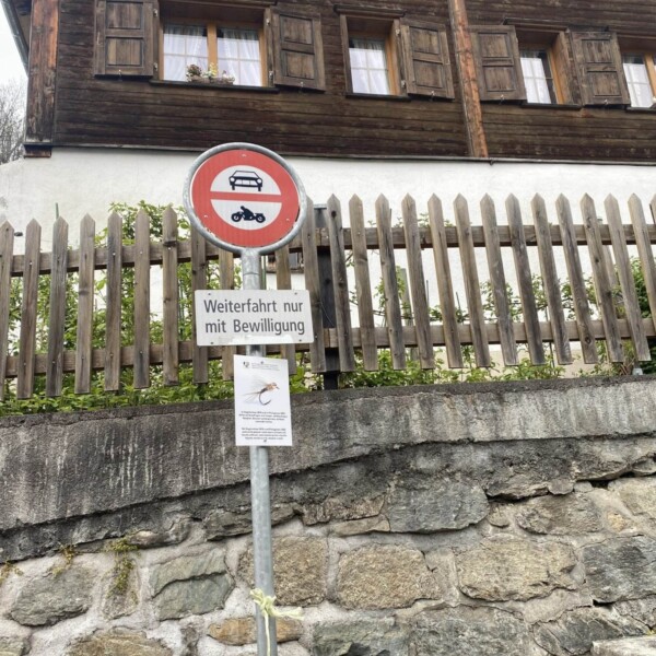 Ein deutsches Verkehrsschild, das Autos und Motorräder verbietet, steht neben einer Steinmauer und einem Holzhaus in der Nähe des Plävigginersees. Unter dem Schild steht ein Hinweis: „Weiterfahrt nur mit Bewilligung“.