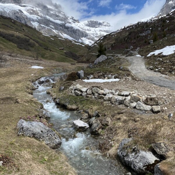 Der Schanielabach fließt durch ein felsiges, grasbewachsenes Tal mit schneebedeckten Flecken, umgeben von Bergen unter einem teilweise bewölkten blauen Himmel. Ein Feldweg folgt dem ruhigen Wasser des Schanielabachs.