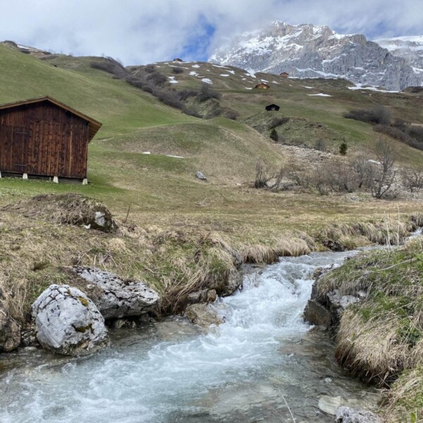 Eine Holzhütte steht auf einem grasbewachsenen Hügel in der Nähe des fließenden Schanielabachs, im Hintergrund sind unter einem leicht bewölkten Himmel teilweise schneebedeckte felsige Berge zu sehen.