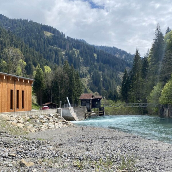 Ein modernes Gebäude mit Holzakzenten steht am schnell fließenden Schanielabach in einem bewaldeten Gebirgstal, mit einer Hängebrücke und einer alten Holzkonstruktion im Hintergrund unter einem teilweise bewölkten Himmel.