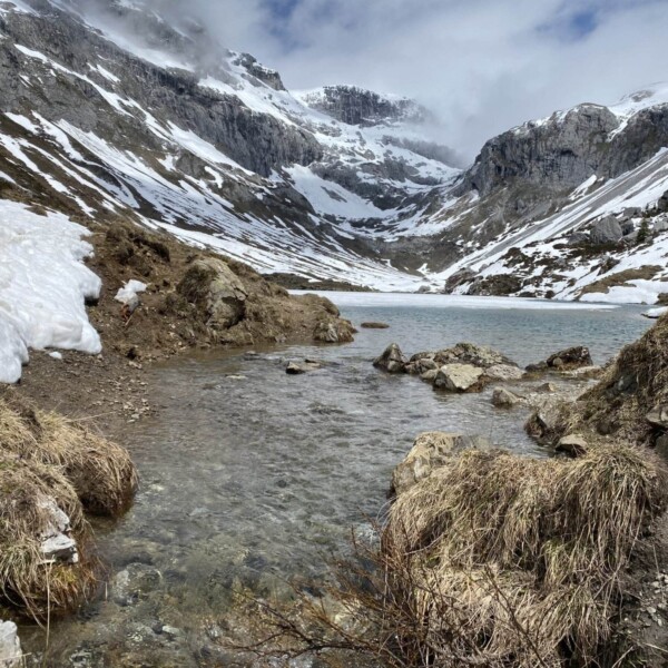 Ein Gebirgstal mit schmelzenden Schneeflecken, felsigem Gelände und dem klaren Schanielabach, der zu einem kleinen See führt, umgeben von schroffen, schneebedeckten Gipfeln unter einem bewölkten Himmel.