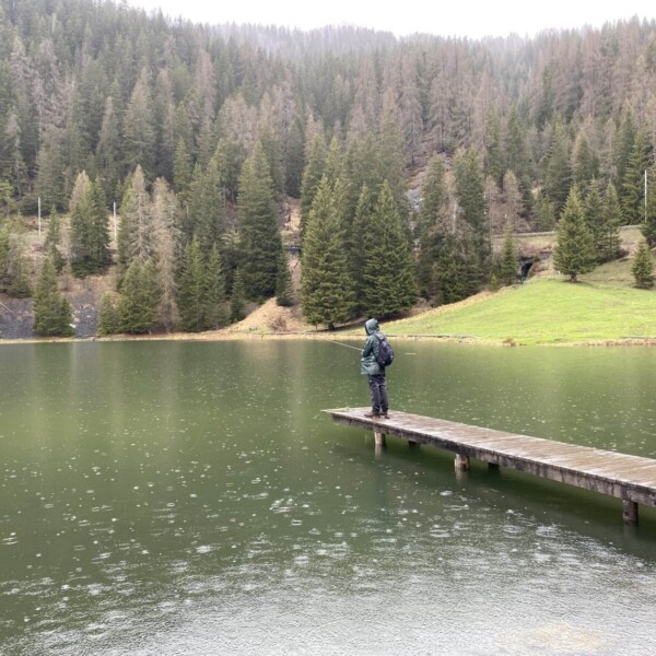 Eine Person im Regenmantel steht auf einem Holzsteg über dem ruhigen Wasser des Davos Laret, umgeben von Tannen und bewaldeten Hügeln unter einem bewölkten Himmel. Regentropfen plätschern über die Oberfläche des Sees.