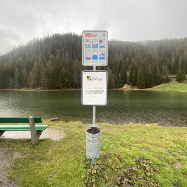 Am Schwarzsee bei Davos Laret steht ein Schild mit Richtlinien und einem Davos-Logo. Am grasbewachsenen Ufer stehen eine grüne Bank und ein Mülleimer, im Hintergrund ein bewaldeter Hügel unter einem bewölkten Himmel.