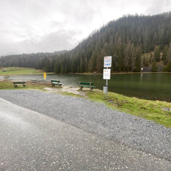 Ein kleiner See, Davos Laret, umgeben von bewaldeten Hügeln unter einem bewölkten Himmel. Drei grüne Bänke stehen am Wasser, neben einem Wegweiser an einem Schotterweg, während sich eine nasse Straße um diese ruhige Ecke von Davos schlängelt.