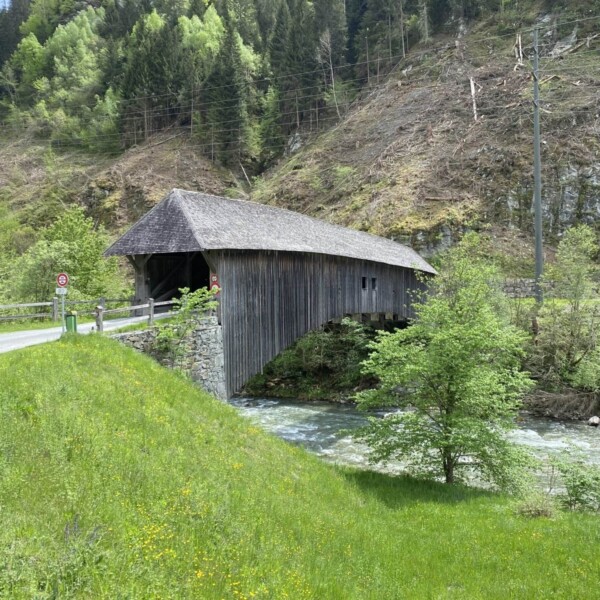 Eine überdachte Holzbrücke überquert einen Fluss in einer üppigen, grünen Berglandschaft, umgeben von Bäumen und Gras. Eine schmale Straße führt zur Brücke. In der Nähe ist ein Geschwindigkeitsbegrenzungsschild zu sehen.