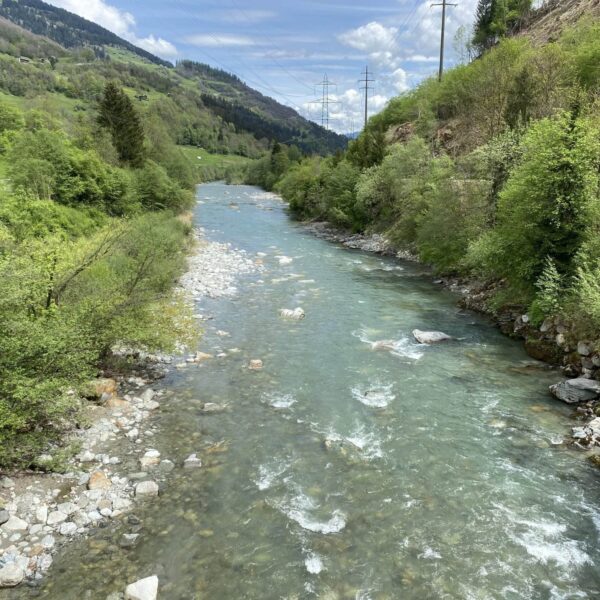 Ein klarer, steiniger Fluss fließt durch ein üppig grünes Tal mit Hügeln und Wäldern auf beiden Seiten; im Hintergrund erheben sich Berge unter einem teilweise bewölkten Himmel.