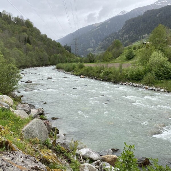 Ein schnell fließender Fluss, gesäumt von Felsen und Grün, schlängelt sich unter einem bewölkten Himmel durch ein üppiges, gebirgiges Tal. In der Ferne sind schneebedeckte Gipfel und Stromleitungen sichtbar.