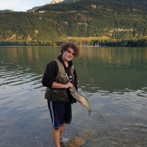 Ein junger Mensch steht im seichten Wasser eines Sees und hält einen Fisch in der Hand. Er trägt ein Unterhemd, Shorts und Slipper. Im Hintergrund erheben sich grüne, baumbedeckte Hügel und Berge unter einem teilweise bewölkten Himmel.