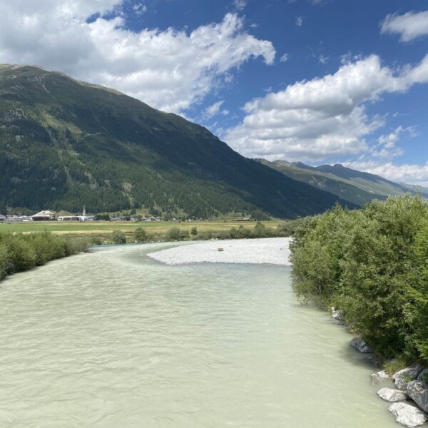 Ein breiter, hellgrüner Fluss fließt durch üppiges Grün mit Bergen im Hintergrund unter einem teilweise bewölkten blauen Himmel. In der Nähe des Flussufers sind Kiesbänke und entfernte Gebäude zu sehen.