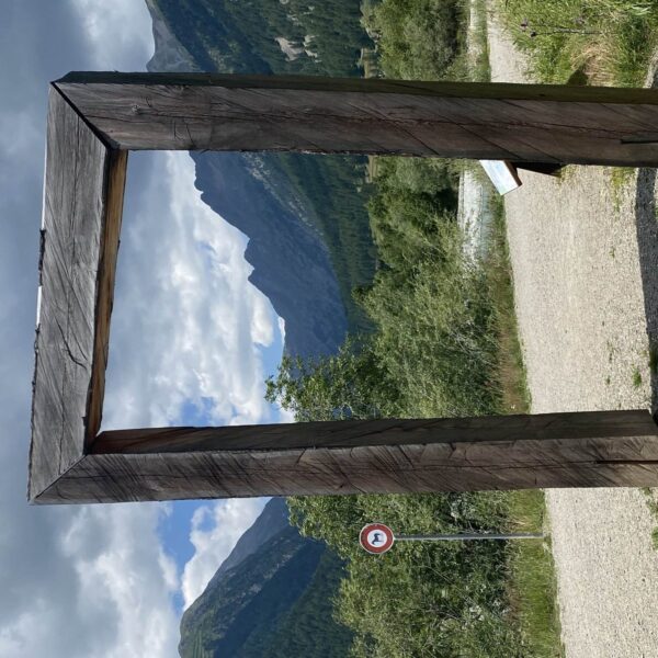 Ein großer rechteckiger Holzrahmen steht auf einem Schotterweg mit Bergen, grünen Bäumen und bewölktem Himmel im Hintergrund. Links vom Rahmen ist ein rundes Verkehrsschild mit rotem Rand zu sehen.